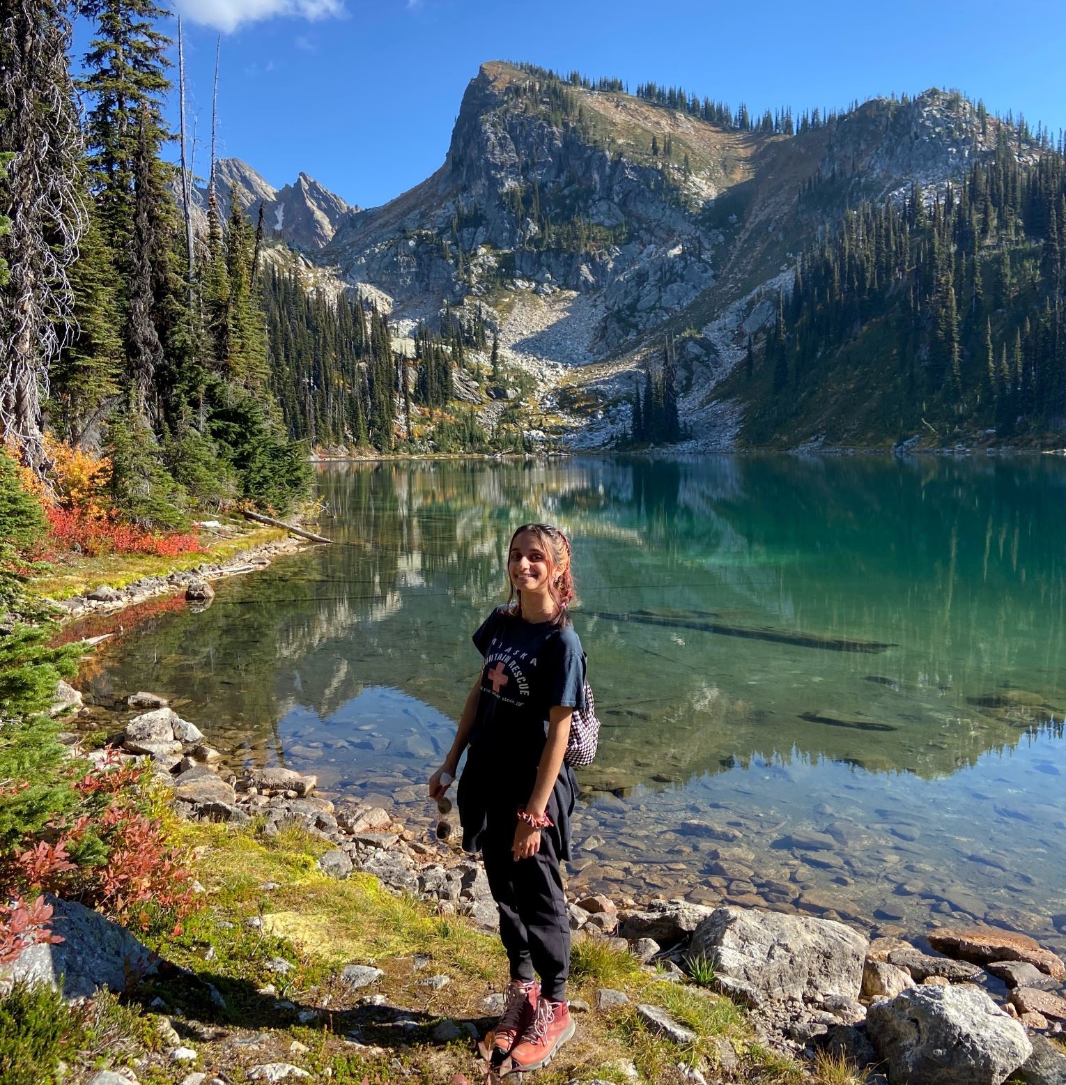 A woman on a hike, standing on the lakes edge with mountains in the background