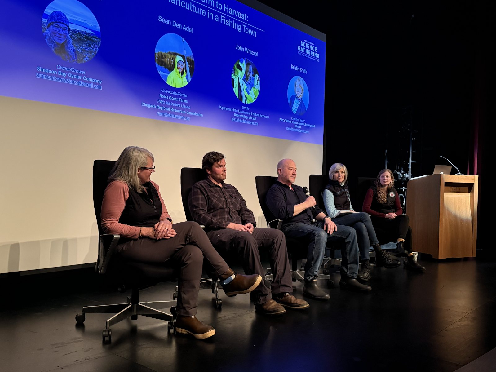 Panelists sit on a stage in front of a PowerPoint presentation.