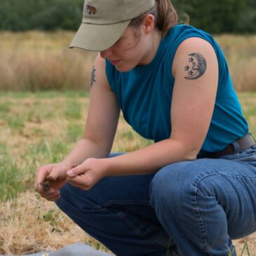 THis photo shows a young woman crouching in a meadow with trees in the background looking at dirt and plants.