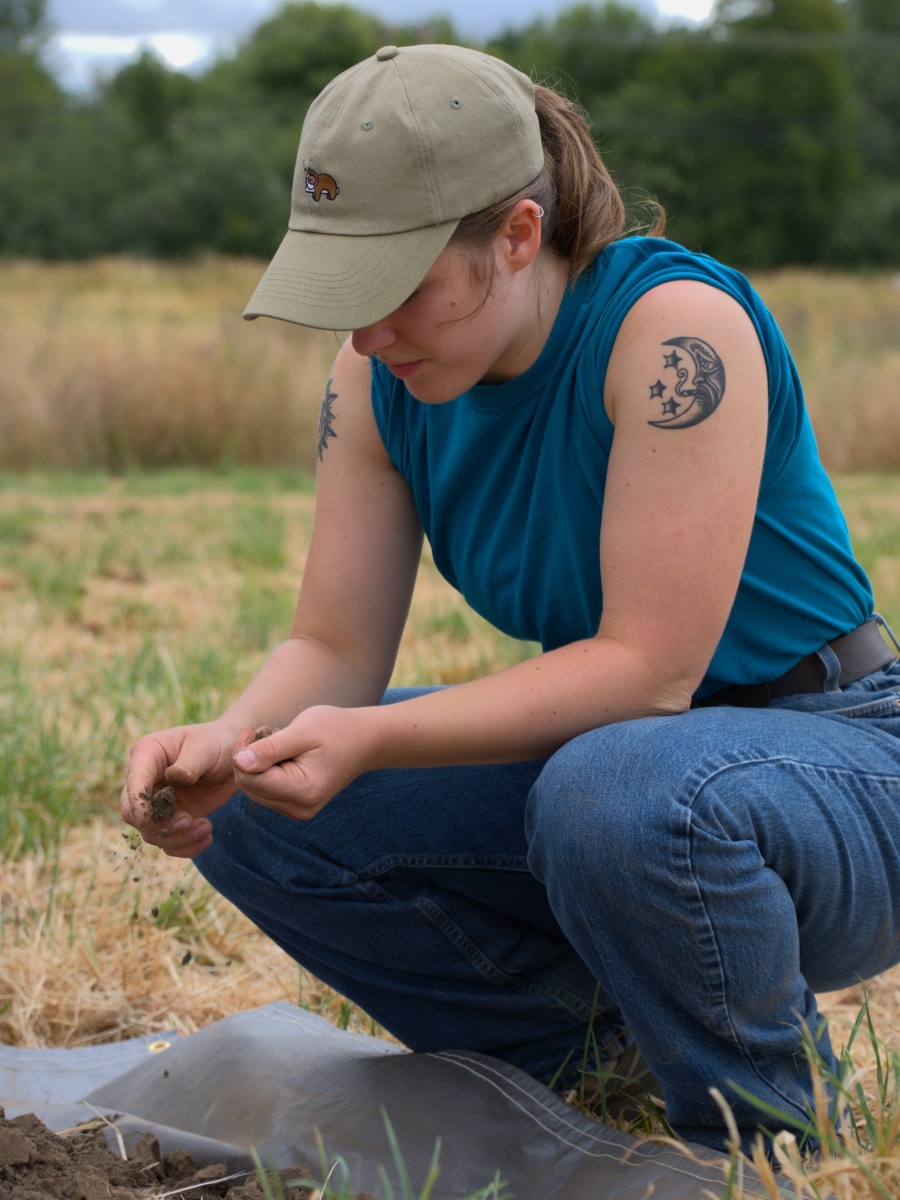 THis photo shows a young woman crouching in a meadow with trees in the background looking at dirt and plants.