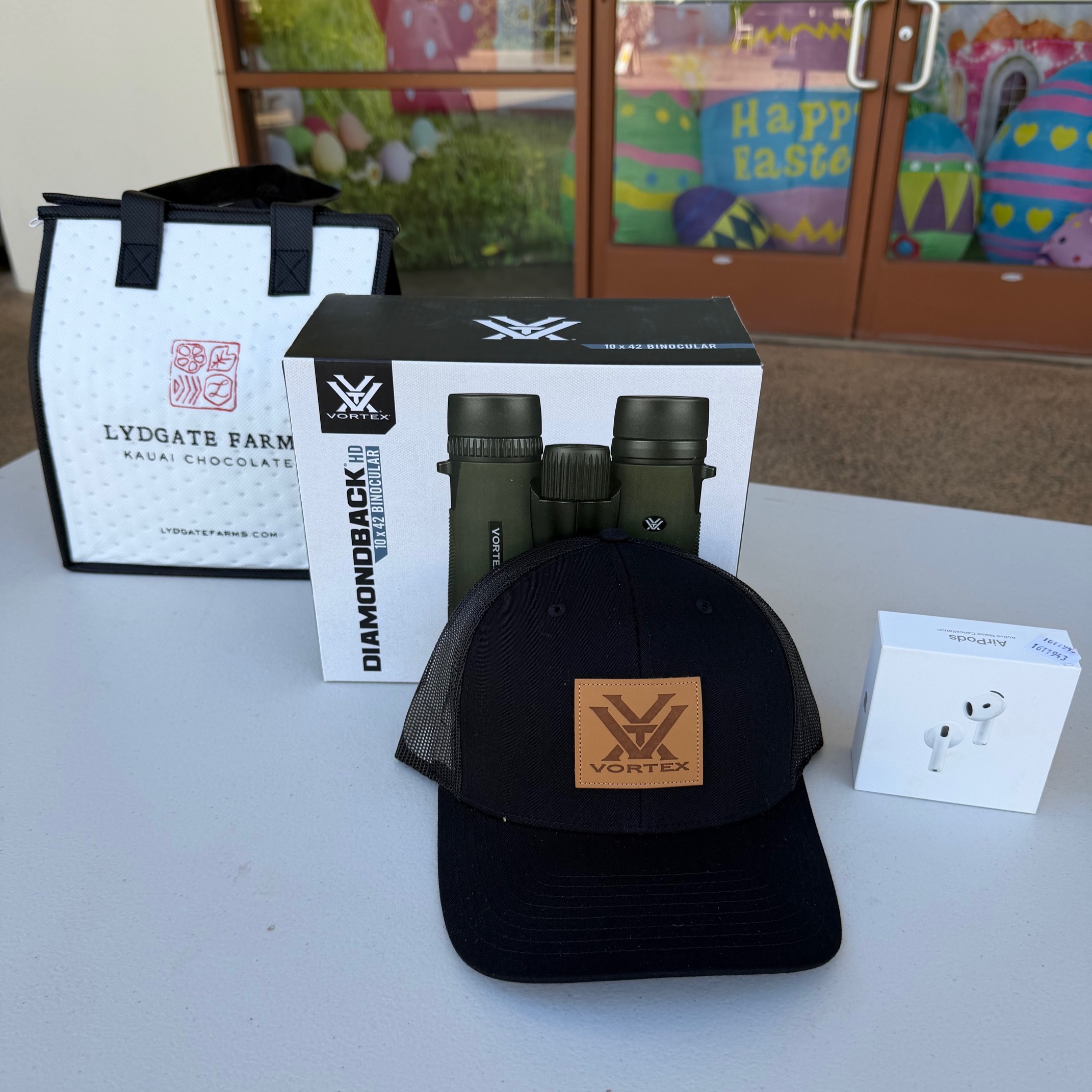 The image shows a bag of Lydgate chocolates, Apple Airpods, a baseball cap, and Vortex binoculars.