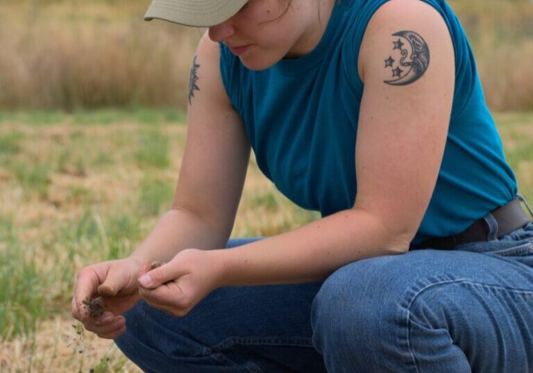 THis photo shows a young woman crouching in a meadow with trees in the background looking at dirt and plants.