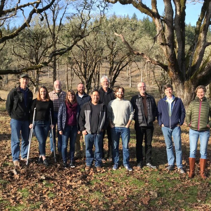 A group of people standing together, looking at the camera wth trees on on a hillside behind them in fall
